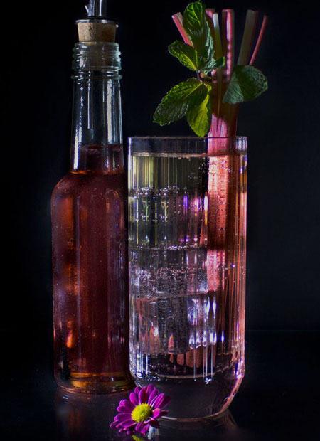 Red syrup in a bottle and long drink glass with rhubarb stalk.